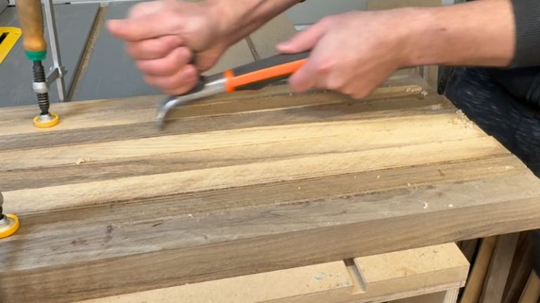 Close-up of a cabinet scraper removing dried glue squeeze-out from the walnut panel.